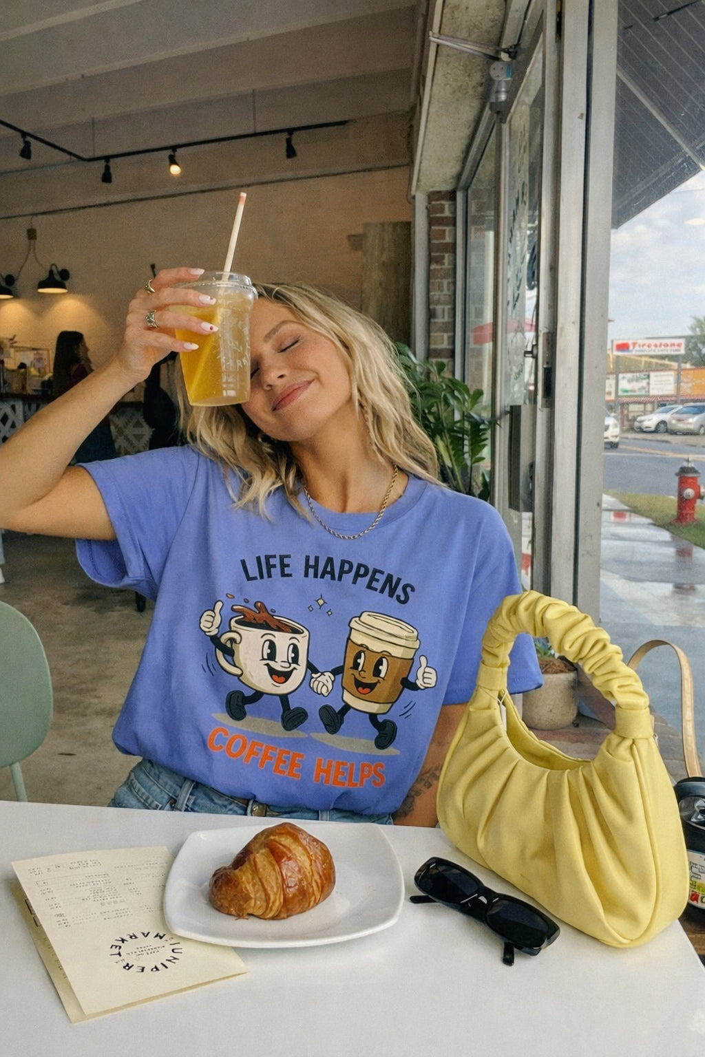 Young woman in a cafe holding iced drink wearing a blue Life Happens Coffee Helps graphic t-shirt with retro coffee cup design in natural iPhone-style photo