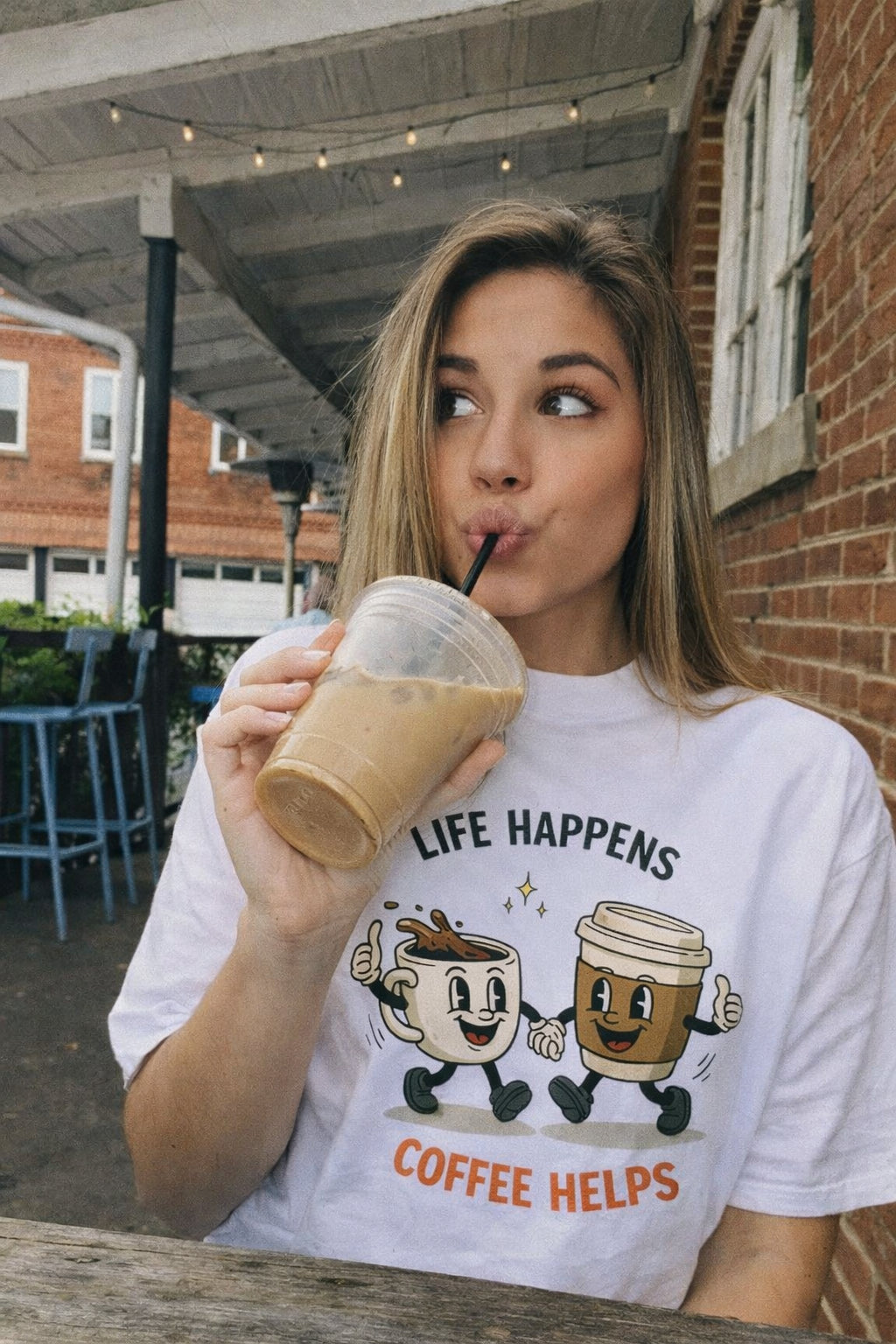 Young woman drinking iced coffee wearing a white Life Happens Coffee Helps graphic t-shirt in a natural outdoor lifestyle photo