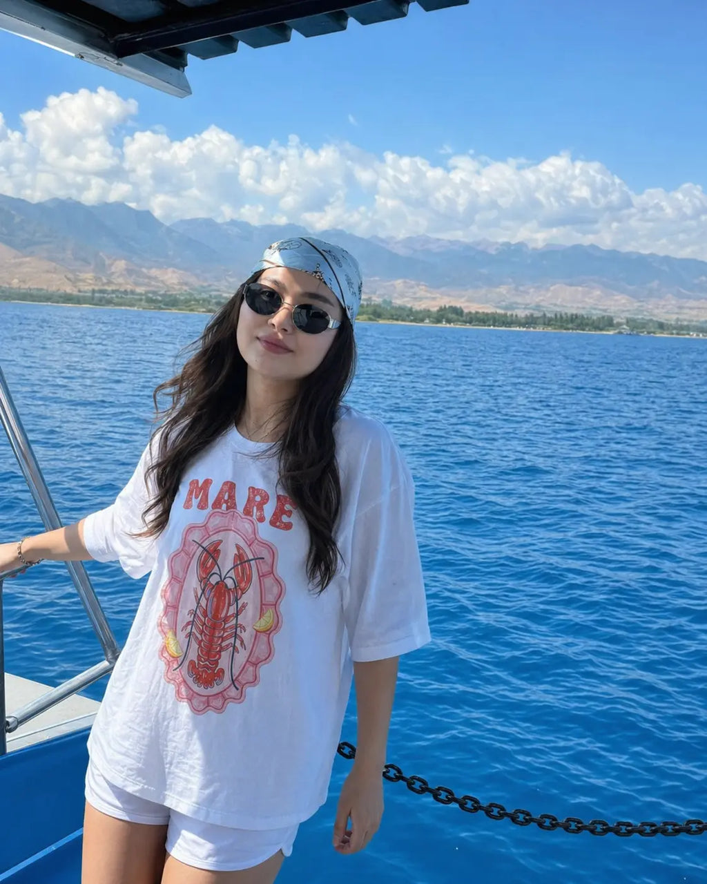 Woman wearing Mare lobster graphic tee in white seated on boat with ocean background — coastal graphic tee for women