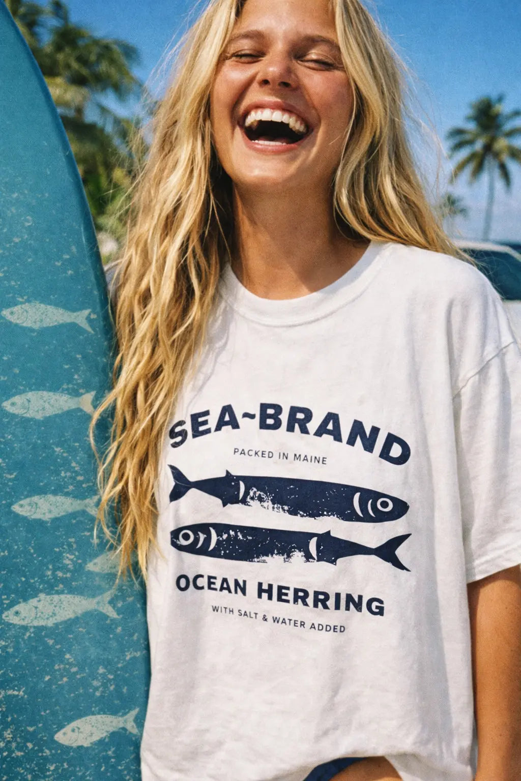 Close-up of a woman wearing a Sea Brand Ocean Herring graphic T-shirt on the beach next to a blue surfboard