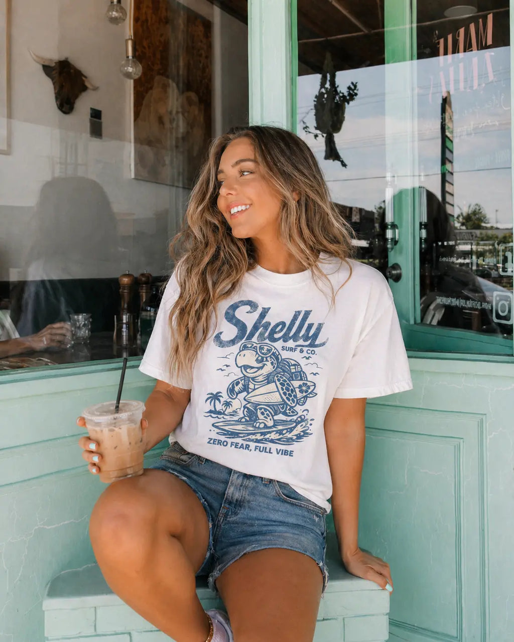Woman holding iced coffee in Shelly Surf sea turtle graphic tee — coastal summer t-shirt for women white