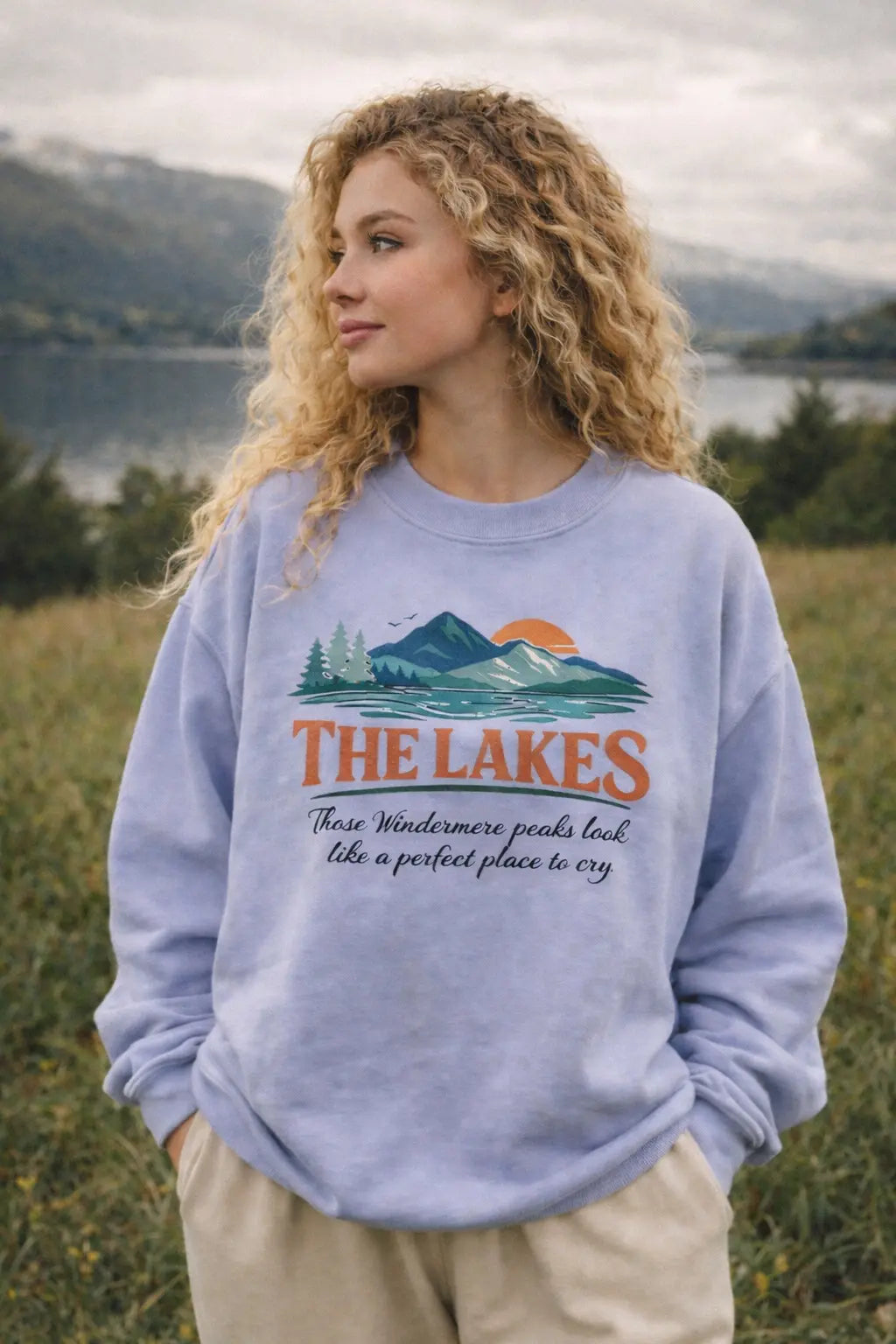 Woman wearing The Lakes crewneck sweatshirt in lavender — vintage mountain and lake graphic with "Those Windermere peaks look like a perfect place to cry" — standing in a meadow with a lake and mountains in the background
