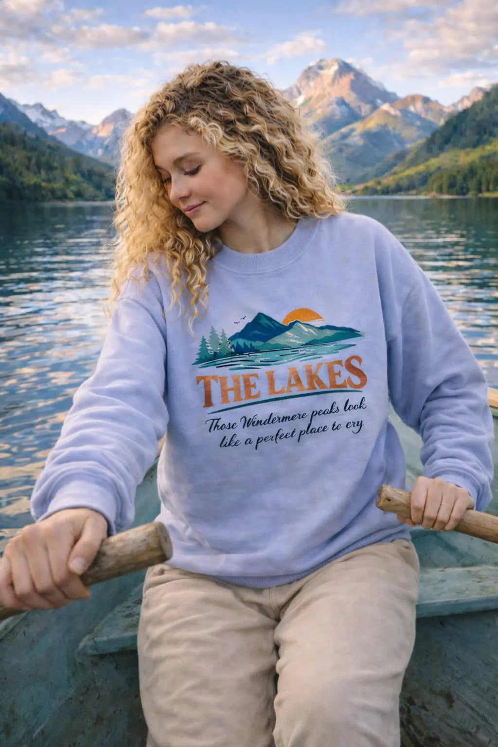 Woman rowing a boat on a lake wearing The Lakes crewneck sweatshirt — lavender vintage graphic pullover with mountain and pine silhouette design, snow-capped peaks in the background