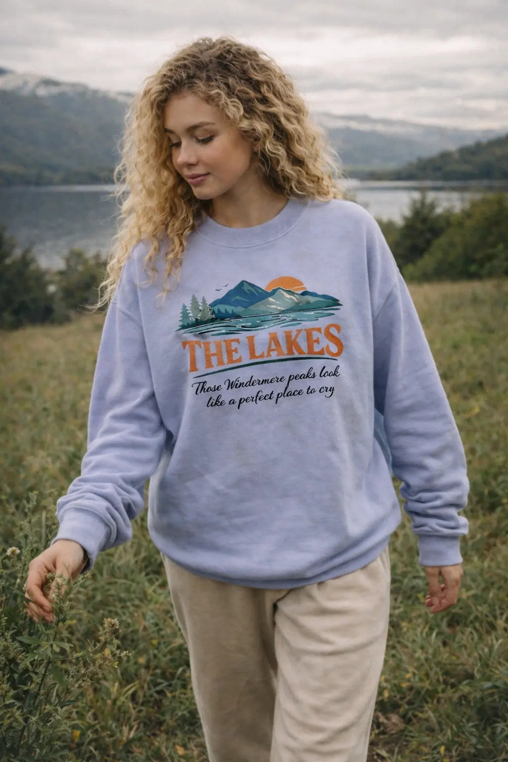 The Lakes vintage graphic pullover in lavender worn by a woman walking through a field near a lake — retro mountain illustration with pine trees and sunset design
