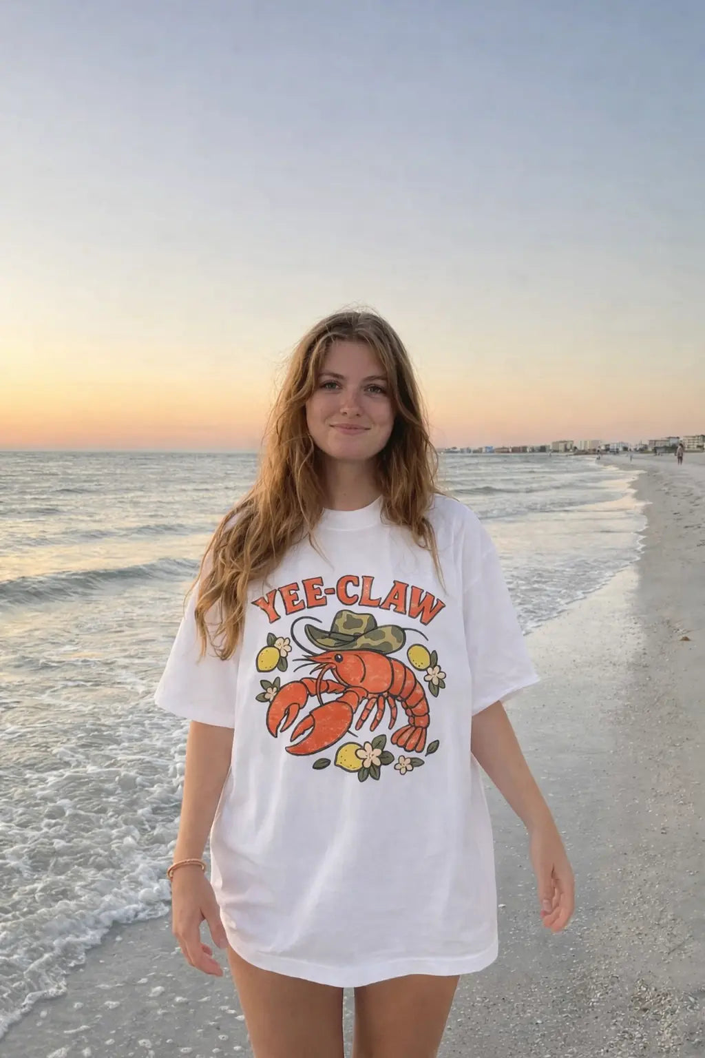 Woman wearing a Yee Claw lobster graphic T-shirt walking on the beach at sunset by the ocean